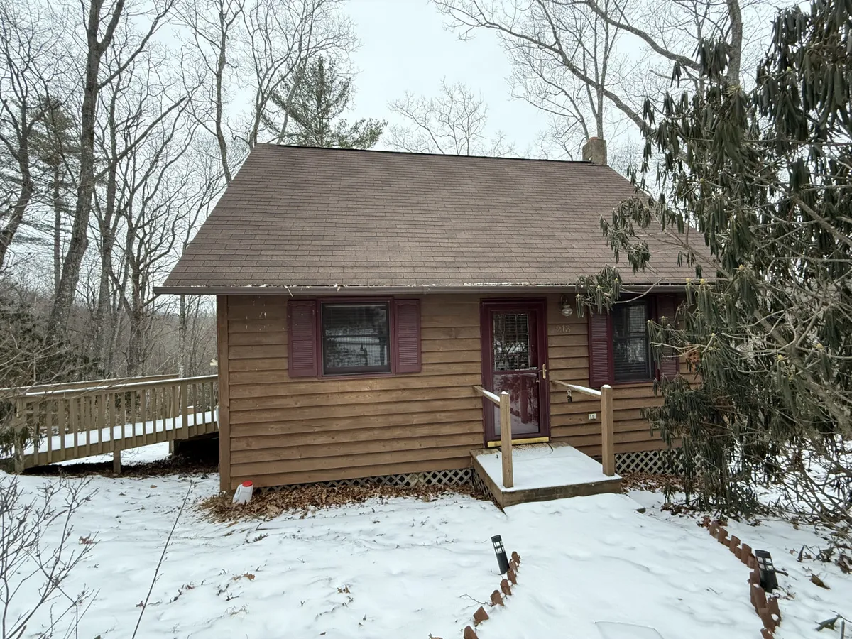 This image showcases the cabin's wooden exterior framed by a snow-covered path, with hints of the inviting porch and picturesque natural surroundings. The setting suggests a peaceful, private retreat nestled in the mountains, perfect for couples seeking a romantic escape.