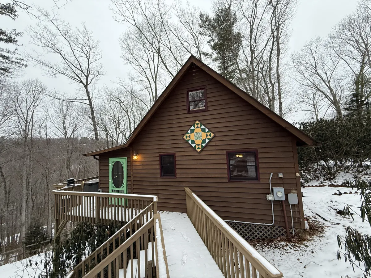The hero image showcases the inviting exterior of a log cabin nestled in the North Carolina mountains. A welcoming wooden porch with a distinctive green door and a vibrant, colorful quilt square sets a cozy, rustic tone. The natural surroundings hint at the tranquility of the setting, perfect for relaxing or gathering with friends by the fire.