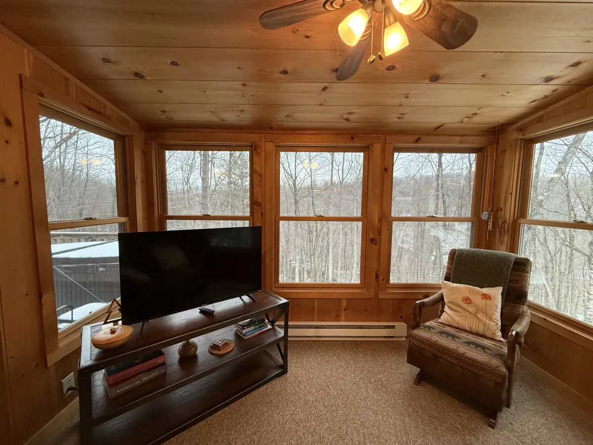 A cozy interior space featuring large windows that let in abundant natural light, with wooden walls contributing to a warm, inviting atmosphere. A comfortable chair is positioned to take in the views, and a TV stand adds to the relaxed feel—suggesting this is a common area where guests can gather, unwind, or enjoy quiet moments while observing nature outside.