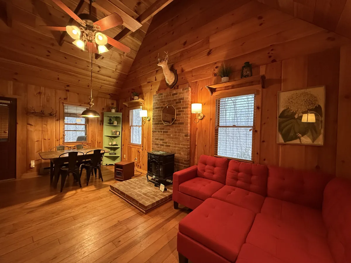 A warm, spacious living room featuring a red sectional sofa and a rustic brick fireplace. The room includes a wooden dining table, vintage decorative elements like a deer head and classic decor items, and is set against a backdrop of timber walls and ceiling that create a cozy cabin vibe. The space is clearly designed for gathering, relaxing, and enjoying fireside moments as a group.