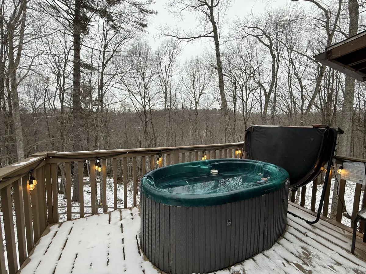 This tranquil exterior shot captures the hot tub on the deck with a view toward the forested surroundings. The deck railing lines the space, and the hot tub sits ready for use. The image conveys a sense of peaceful seclusion, inviting viewers to imagine soaking while enjoying the sunset or watching snow fall through the trees.