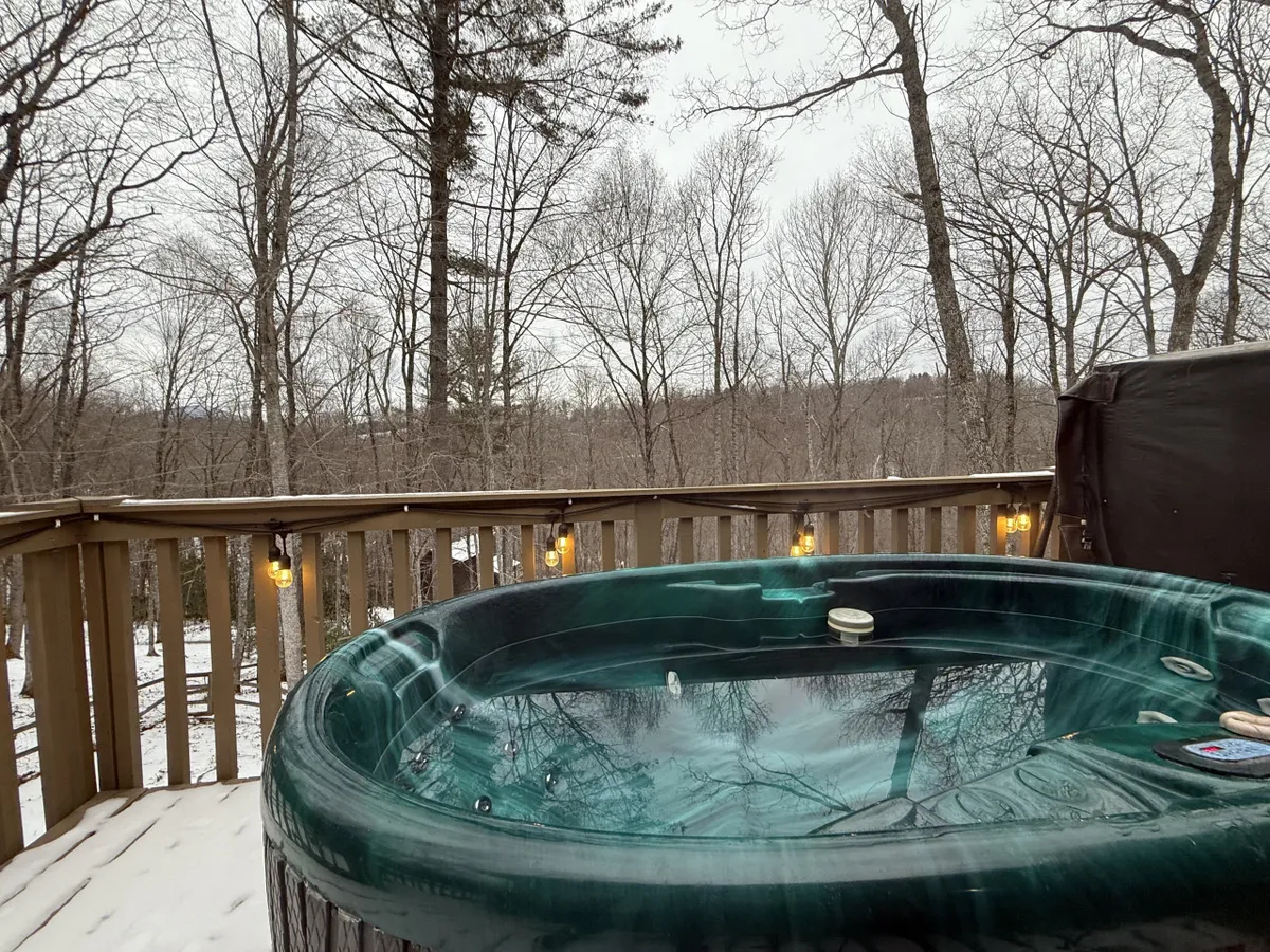 This exterior image showcases the inviting hot tub nestled on the spacious wooden deck, surrounded by towering pines and glowing hanging lights. The scene feels serene, with the hot tub promising relaxation amid nature, and hints of snow adding a magical touch. The railing frames the deck, and the setting is clearly designed for peaceful moments and socializing under the sky.