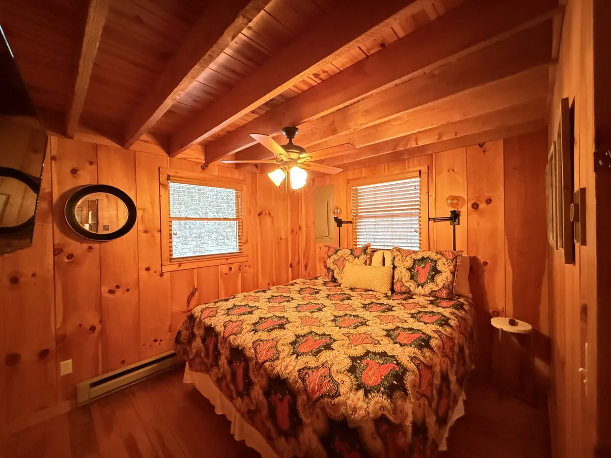 This image showcases a cozy queen bed adorned with patterned bedding, surrounded by warm wooden walls and soft natural light streaming through a window. The bedroom is enhanced with a ceiling fan and a decorative mirror, evoking a tranquil and inviting atmosphere ideal for restful nights.