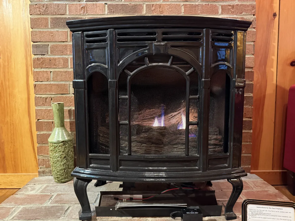 A cozy interior scene focused on a black fireplace with a green vase on the hearth, set against a brick wall. The space feels intimate and perfect for gathering, with a sense of warmth that echoes the idea of stories and marshmallow roasting around a fire—evoking the firepit experience described in the section.