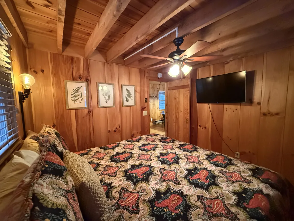 This image presents another angle of a bedroom with a plush bed dressed in detailed bedding, set against wood paneling and accented by framed botanical prints and soft overhead lighting. A TV on the wall adds a touch of modern comfort, while the overall ambiance remains warm and intimate.