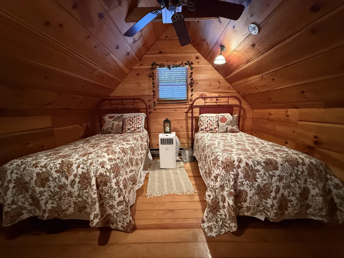 This interior shot displays two twin beds tucked into a cozy, loft-like space with wooden wall paneling, decorative pillows, and a small window with blinds. Soft, inviting lighting and a ceiling fan complete the scene, suggesting a comfortable and welcoming environment for children or friends.