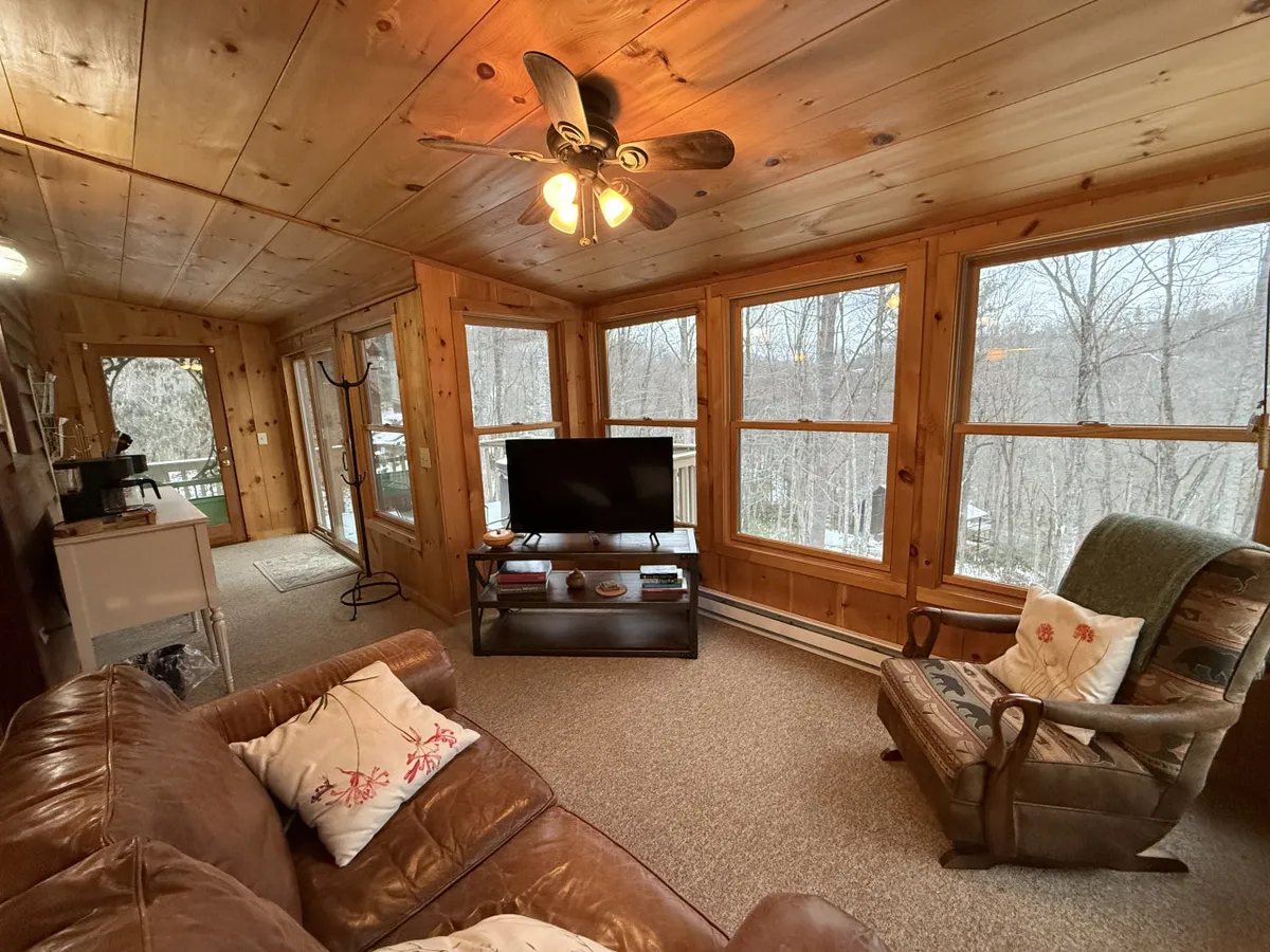 A comfortable leather sofa sits beneath exposed wooden ceilings and walls, facing a TV unit adorned with tasteful decor. Large windows let in natural light and provide woodland views, connecting the cabin’s interior with the outdoors. The space feels inviting and is ideal for relaxing, watching movies, or enjoying the view with a morning coffee.