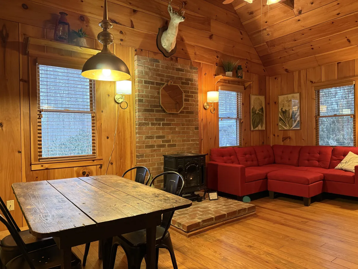 A warm, inviting interior scene showcasing a wooden dining table perfect for family meals, a red sectional sofa for group relaxation, and a prominent brick fireplace—an ideal spot for gathering after a day of adventures. The rustic decor, including wall art and an antler mount, emphasizes the cozy, communal spirit of the lodge.